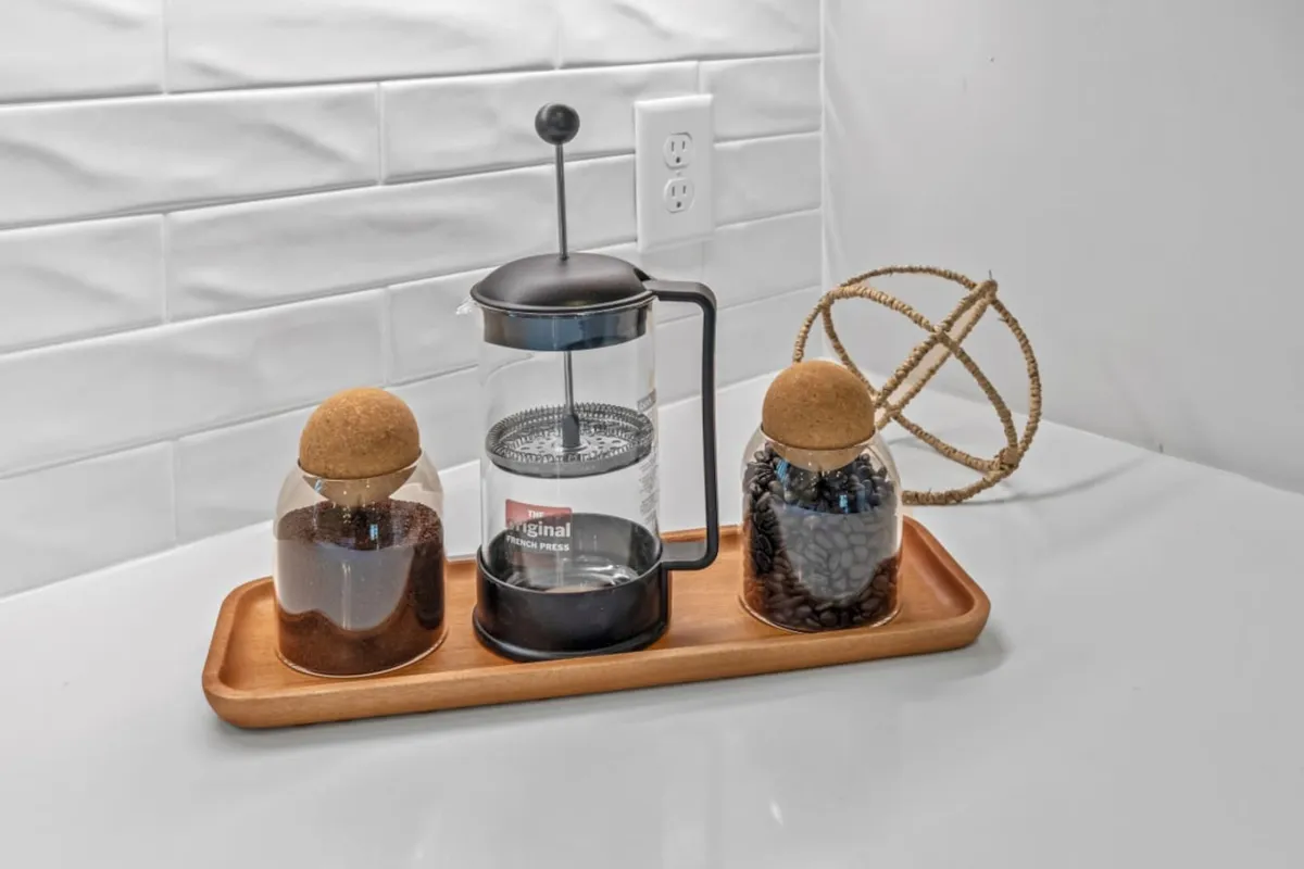 A cozy kitchen scene featuring a French press, glass jars with cork lids, and a wooden tray arranged neatly on the countertop. The inviting setup suggests a communal gathering spot for morning coffee or casual conversations, with soft natural light enhancing the welcoming atmosphere.
