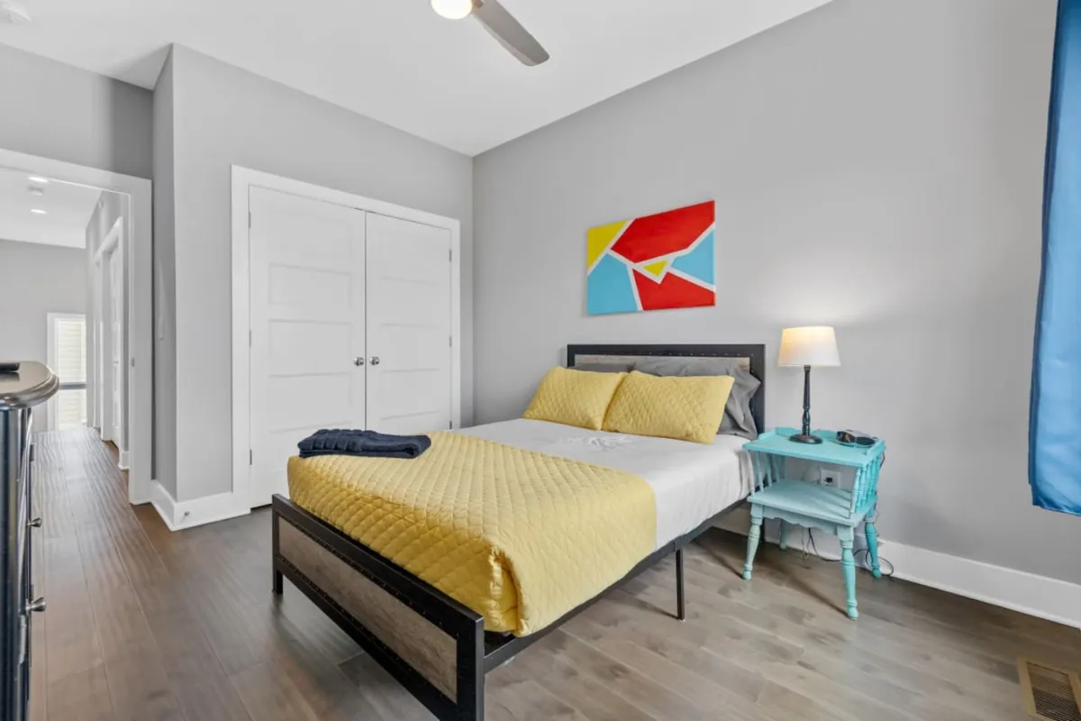 A comfortable bedroom with a bed dressed in yellow bedding, colorful artwork on the wall, and a blue nightstand with a lamp. The space is filled with natural light, creating a bright and uplifting atmosphere ideal for relaxing after a productive day.