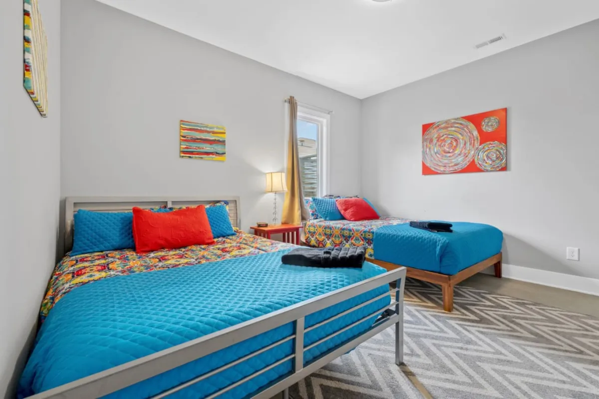 A vibrant, cozy bedroom with colorful bedding, lively wall art, and a glowing table lamp. The space feels inviting, perfect for friends to enjoy late-night conversations or unwind after an evening out.