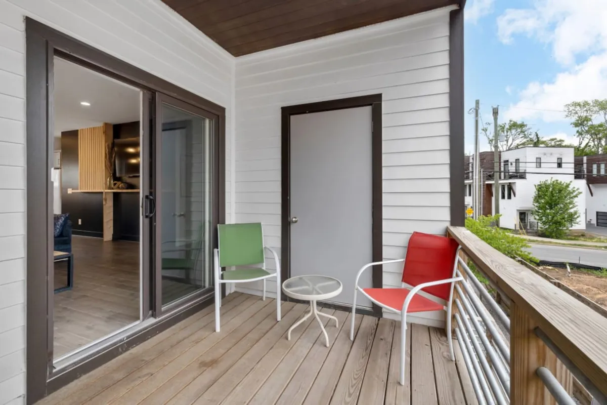 This image showcases an outdoor balcony featuring a red and green chair, a small table, and a glimpse of the street below. The mood is casual and welcoming, suited for relaxing, socializing, or enjoying a beverage while taking in the neighborhood views.