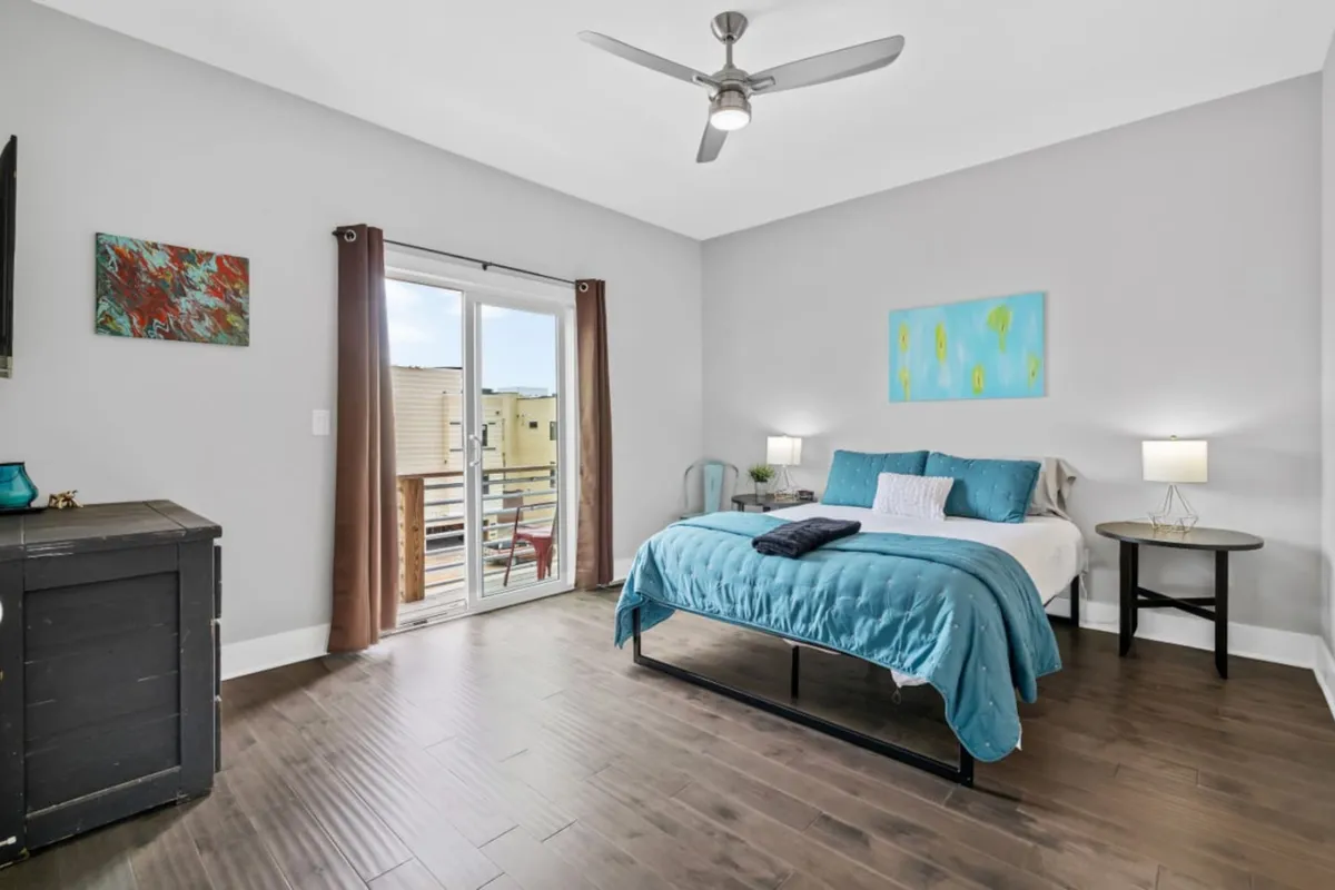A serene bedroom with blue bedding, sliding glass doors leading to a private balcony, tasteful artwork, and nightstands with lamps. Natural light streams in, creating a tranquil and restful ambiance.