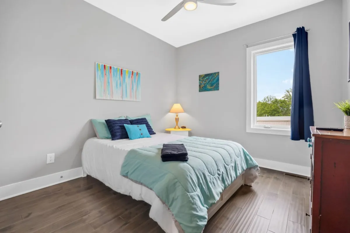 A tranquil bedroom is shown with a bed dressed in teal bedding, artwork on the walls, a bedside lamp, and a window letting in natural light. The setting suggests restful moments, reading, or quiet contemplation by the window.