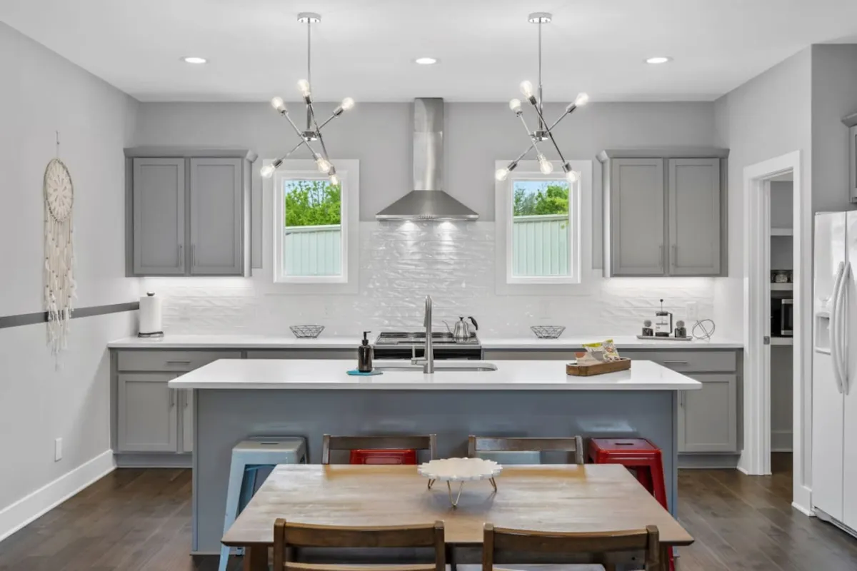 A spacious chef’s kitchen featuring a central island, a large dining table with seating for the whole family, modern light fixtures, and big windows that allow natural light to pour in. The scene is set up for cooking, dining, socializing, and entertaining, capturing the heart of family gatherings and shared meals.